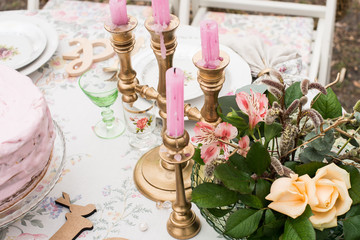 Pink candles in a candlestick on a table with a few roses and greenery in a glass jar