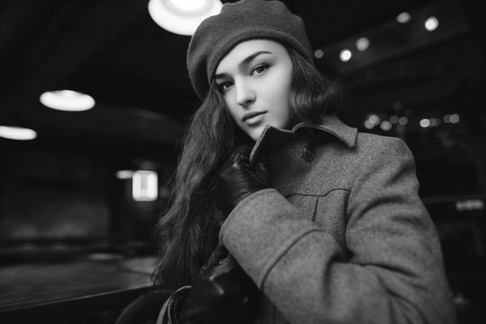 Indoor monochrome photo of young pretty fashionable girl with long hair. Model wearing stylish woolen coat and beret. Waist up. Beautiful lights on background. Female fashion concept
