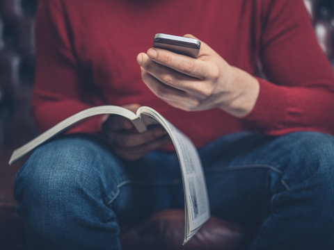 Man on sofa reading booklet and using smartphone