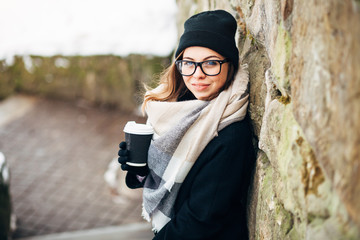 Portrait of a beautiful girl in black glasses in a hat and coat and gloves with scarf in the winter park holding warm coffee