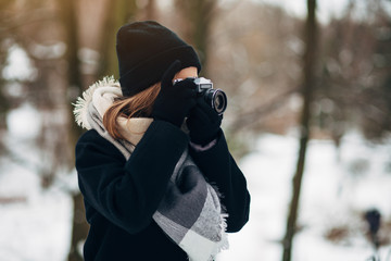 Girl photographer in black glasses in a hat and coat and gloves with scarf in the winter park makes photo on old retro film camera