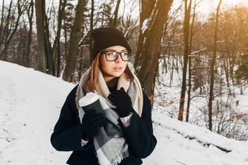 Portrait of a beautiful girl in black glasses in a black hat and coat and gloves with scarf in the winter park holding warm coffee