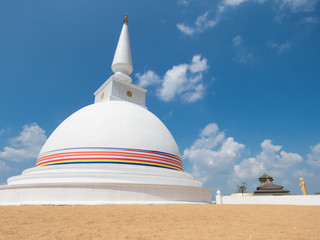 White Stupa Dagabo on sand wrapped with Buddhist flag and blue sky with clouds and Temple palace in...