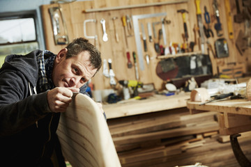 Man standing in a workshop inspecting closely the edge of a wooden surfboard. 