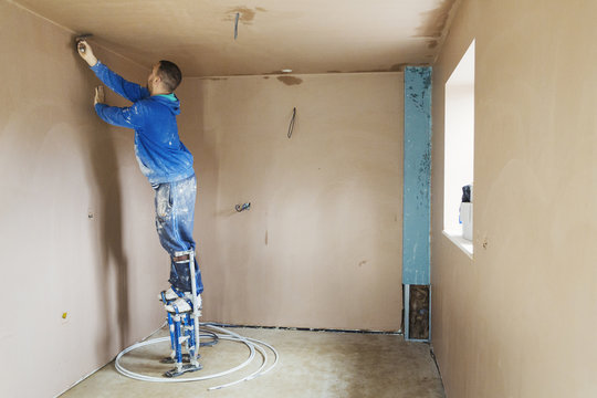 A plasterer wearing stilts smoothing fresh plaster high up on the walls of a house under construction. 