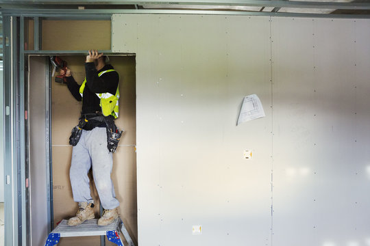 A carpenter in work clothes on a construction site, using an electric drill. 