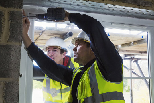Two workmen on a construction site, builder in hard hat using an electric drill on a window frame. 