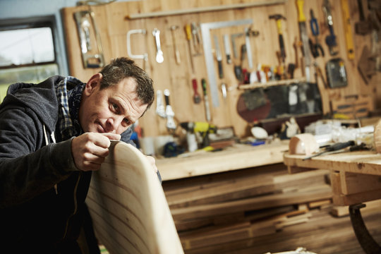 Man standing in a workshop inspecting closely the edge of a wooden surfboard.  - Powered by Adobe