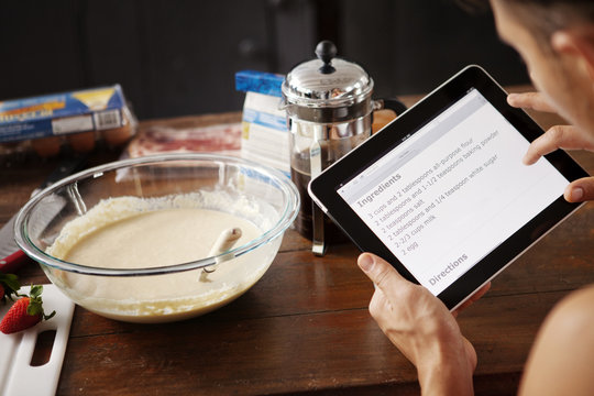 Man baking pancakes with recipe on digital tablet