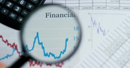 A hand with a magnifying glass passes over a financial newspaper and financial bag with a business plan. The lens magnifies the word "Financial". Concept: economy, finance, crisis, business plan, - Powered by Adobe