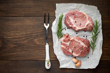 Two pieces of raw pork meat on white parchment with herb and spices on rustic wooden background.