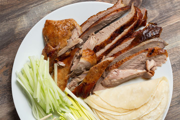 Peking Duck - Chinese roast duck with cucumber pieces, onion, pancakes on white plate close up. Wooden table and top view