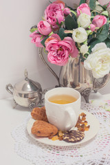 Afternoon tea time party. White cup of green light tea with cakes raisin, chocolate cookies in shabby chic elegant traditional interior on lace napkin with pink flowers. Cookie heart shape. 