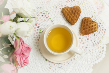 Obraz na płótnie Canvas Happy Valentines day tea time or love party. White cup of green tea with cakes raisin chocolate cookies in shabby chic elegant traditional interior, lace napkin with pink flowers. Cookie heart shape.