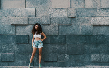 Attractive curly brunette hipster smiling girl in teal jeans shorts in front of street stoned patterned wall background with copy space for your advertising text message or promotional content