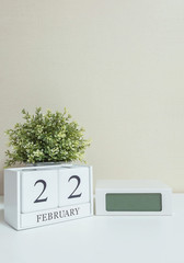 White wooden calendar with black 22 february word with clock and plant on white wood desk and cream wallpaper textured background in selective focus at the calendar