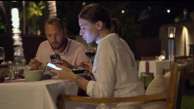 Couple eating meal and using smartphones in restaurant at night

