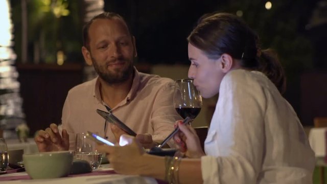 Couple with smartphones talking and eating meal in restaurant at night
