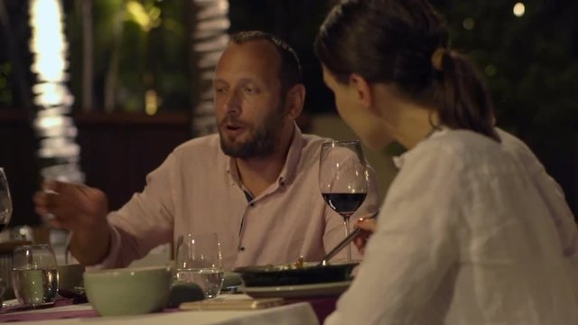 Happy, young couple talking, eating and raising toast with wine in restaurant at night

