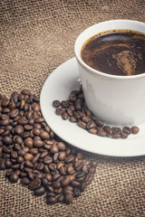 White cup of coffee standing on the burlap sack. Scattered coffee beans are around the cup. Edited as a vintage photo with dark edges. 