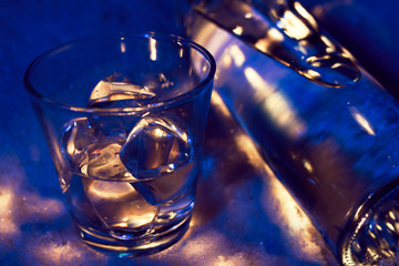 Bottle of vodka in the snow with glass filled with ice cubes, dark blue lit with white highlights 