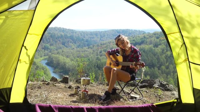Young blonde woman tourist sitting on camping chair on a cliff above the river Berd with guitar and basking in the sun. Russia, Siberia, Salair