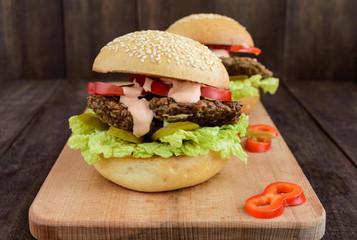 Burgers with liver cutlet, tomatoes, pickles, lettuce, spicy sauce and a soft bun with sesame seeds on a cutting board on a dark wooden background.