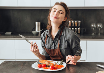 Woman sitting in kitchen while eating fish and tomatoes.