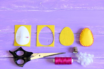 Fototapeta premium How to make a felt Easter egg ornament. Yellow felt Easter egg, cut felt piece in shape of a egg, paper template, scrap, scissors, thread, thimble on a wooden background with copy space for text