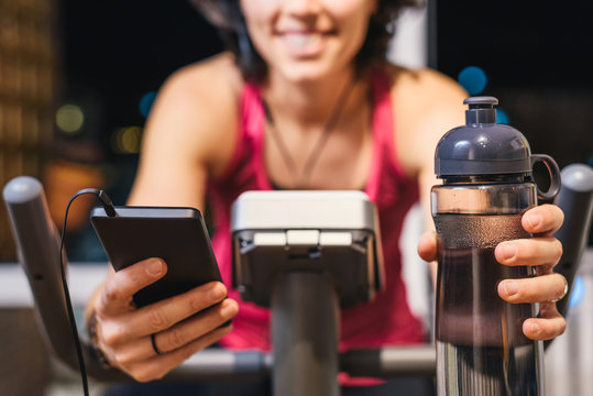 Healthy fit smiling woman training at home on exercise static bike during workout holding phone and bottle of water while listening music with earphones for motivation. Female health weekly habits app