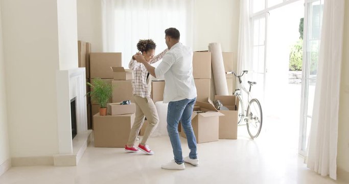 Happy young couple celebrating moving home dancing together in the living room surrounded by packing boxes