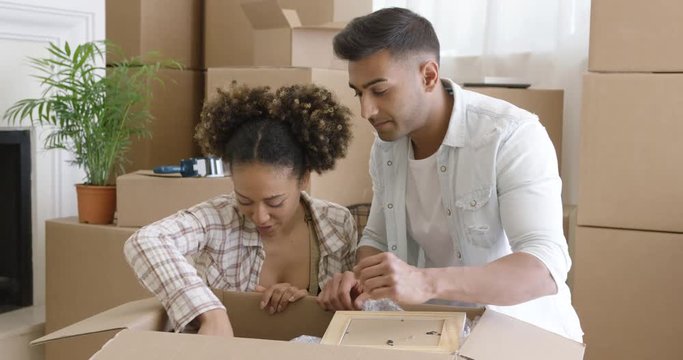 Attractive mixed race couple sitting on the floor with opened box and unpacking their things in new home. Girl holding frame with photo.