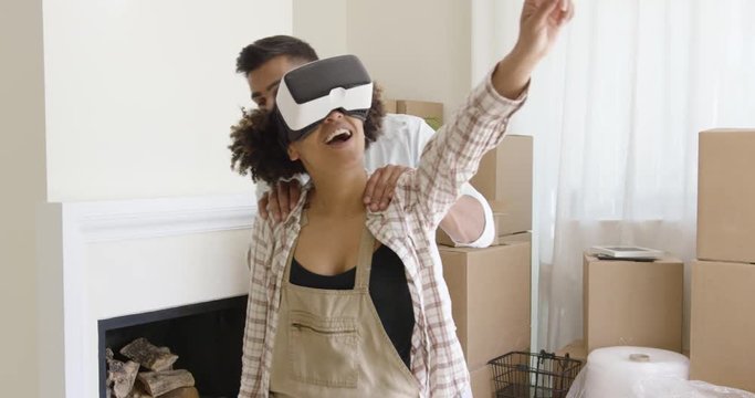 Happy young mixed race couple doing Virtual tour in their fresh bought new apartment. Girl wearing VR goggles and looking around while her partner guides her.
