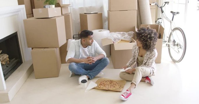 Young couple taking a break from moving house sitting on the floor in front of a pile of boxes enjoying a pizza