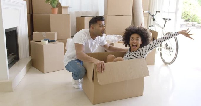 Happy young couple moving house together with the pretty young woman sitting inside a brown cardboard box with her husband crouching alongside smiling