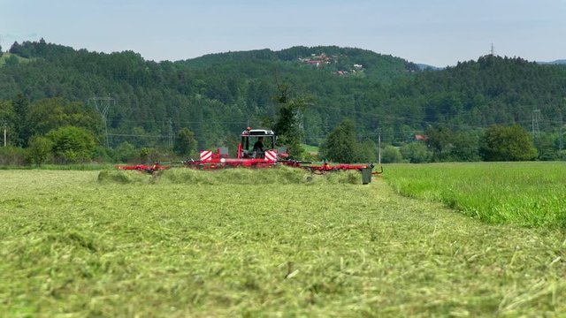 A farmer is driving his big tractor and he is preparing hay on a large field with a huge machinery. It is a beautiful summer day.
