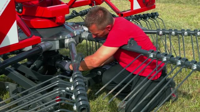 A young man kneels down and he is fixing something on the lower part of the agricultural machinery. He is wearing safety work gloves.
