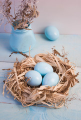 Blue Easter eggs in nest, vase with dry grass on wooden background.