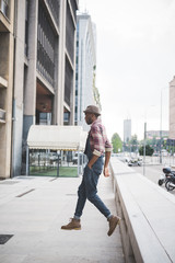 Young handsome afro black man jumping in the street of the city, looking upward surprised, holding his hat on his head - surprised, jumping, having fun concept