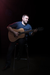 Fototapeta na wymiar Portrait of handsome bearded man on a chair with an acoustic guitar in hand, isolated on a black background
