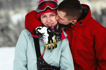 Man in red ski jacket kisses woman's pink cheek and hugs her neck somewhere in snowed forest