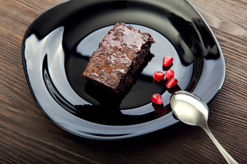 slice of brownie on black plate on a wooden table with a spoon and pomegranate seeds