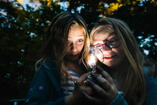Two girls holding and staring at illuminated lightbulb in garden at dusk