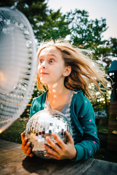 Girl pulling face in front of windy electric fan at garden table