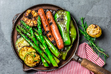 Grilled vegetables and sausages on cast iron grill pan. Gray slate background.