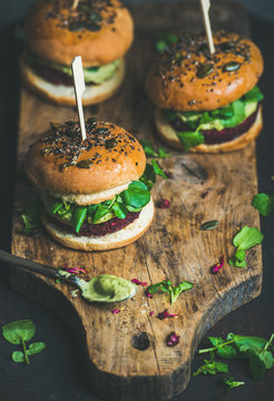 Healthy vegan burgers with beetroot and quinoa patty, arugula, avocado sauce, wholegrain bun on rustic wooden board over dark background, selective focus, copy space. Vegetarian, dieting food concept