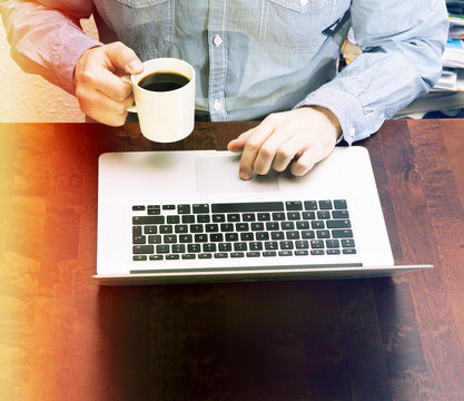 Hipster Doing Work On A Laptop And Holds A Cup Of Coffee. Image Has A Light Leak Effect Applied.