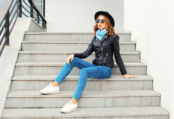 Fashion woman wearing a black rock jacket, hat sitting in city