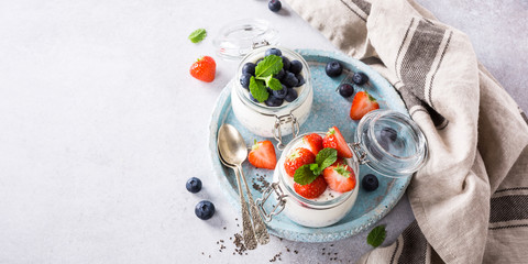 Chia Pudding for breakfast in glass jars garnished with berry in two portions on light gray stone background with a napkin and spoons. Copy space. Top view.