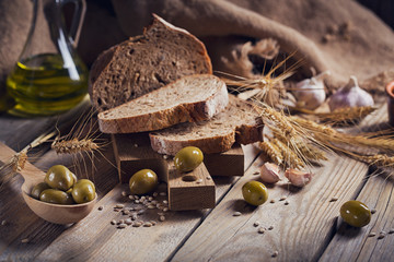 Fresh multigrain crusty bread, green olives and wheat ears on a rustic wooden table.